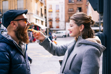 A happy blonde Caucasian girl gives a piece of pizza to a guy with a beard and glasses on a city street