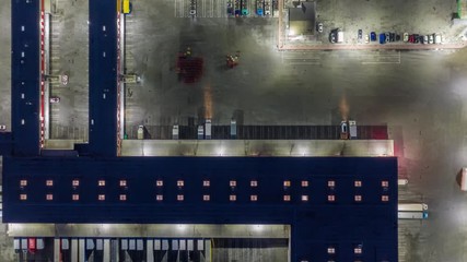 Wall Mural - Aerial top down view of the large logistics park with warehouse, loading hub with semi trucks with cargo trailers standing at the ramps for load/unload goods at night. Hyper lapse - time lapse. Zoom