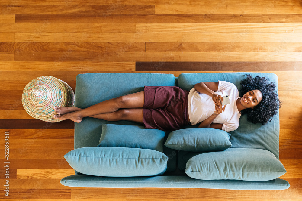 Black woman chatting on smartphone lying on couch at home - obrazy, fototapety, plakaty 