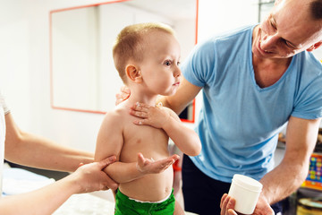 Father and caucasian son two years old child apply skin care cream to his naked body while father and mother are holding kid family at home