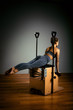 © Georgii - Young girl doing pilates exercises with a reformer bed. Beautiful slim fitness trainer on reformer gray background, low key, art light. Fitness concept
