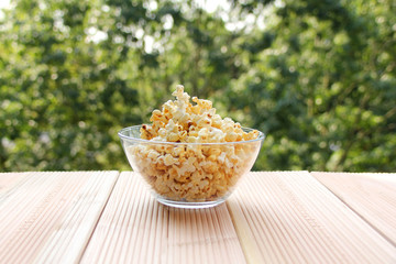 glass cup with popcorn on a light wooden table against a green summer garden, close-up, copy space