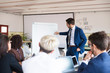 © Mangostar - Confident business trainer drawing on whiteboard. Group of workers sitting at table and listening speaker. Business meeting concept