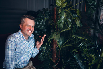 Portrait of senior middle adult man in blue shirt with green plants looking at camera and smile. Home gardening plant.