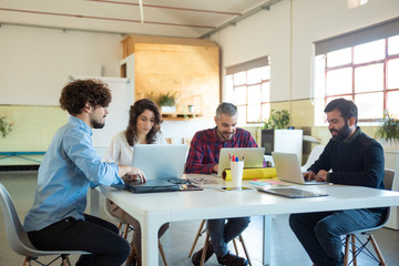 Wall Mural - Cheerful young colleagues discussing project. Smiling male and female coworkers sitting at table with laptops in open space office and talking. Technology, communication concept