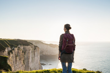 Wall Mural - Young woman traveler with backpack looking at sea in Normandy, France over beautiful cliffs background. Travel, active lifestyle and summer holiday concept