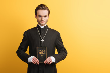 Wall Mural - young, confident catholic priest looking at camera while holding bible isolated on yellow