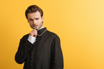 Wall Mural - tense catholic priest looking away while holding cross near lips isolated on yellow