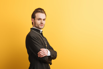 Canvas Print - confident catholic priest with crossed arms looking at camera isolated on yellow