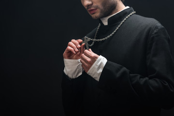 Canvas Print - cropped view of catholic priest looking at silver cross on his necklace isolated on black