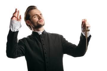 Wall Mural - young catholic priest praying with closed eyes and raised hands while holding golden cross isolated on white