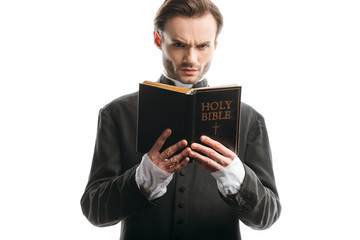 Canvas Print - frowning catholic priest looking at camera while holding holy bible isolated on white