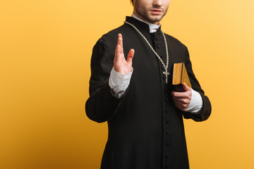 Wall Mural - cropped view of catholic priest showing blessing gesture while holding bible isolated on yellow