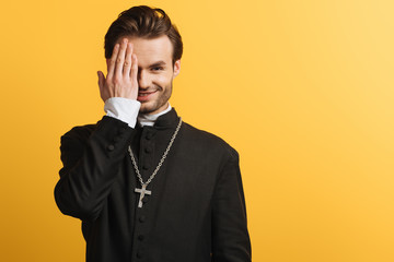 Wall Mural - smiling catholic priest covering eye with hand while looking at camera isolated on yellow