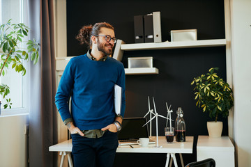 Handsome positive young caucasian hipster standing in his office, holding under armpit and preparing to go on meeting with important clients.