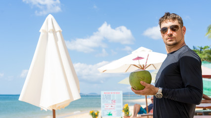 Summer vacation of hansome well shaped young man in sunglasses chilling on the blue sea and drinking coconut .Beautiful beach view with traveler man relaxing on Phu Quoc island, Vietnam. 