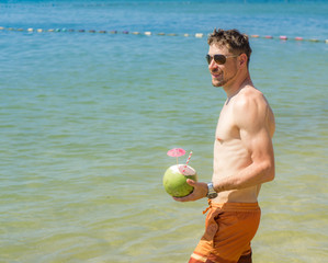 Summer vacation of hansome well shaped young man in sunglasses chilling on the blue sea and drinking coconut .Beautiful beach view with traveler man relaxing on Phu Quoc island, Vietnam. 