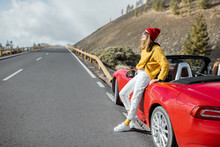 Lifestyle Portrait Of A Carefree Woman Dressed Casually In Yellow Sweater And Red Hat Standing Near The Car On The Roadside, Enjoying Road Trip In The Mountains