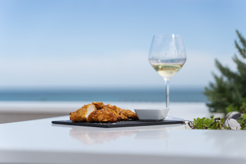 Fried chicken served with sauce in a dark board with a glass of wine in a table in front o f the sea