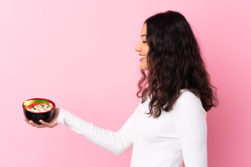 Wall Mural - Mixed race woman holding a bowl full of noodles over isolated pink background with happy expression