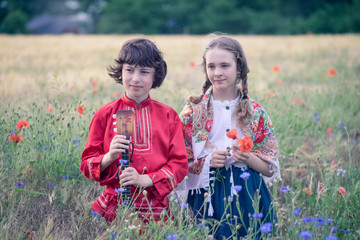 Portrait of a boy and a girl in the field. The girl’s shoulders are covered with a shawl with Russian folk patterns. The boy is wearing a Russian peasant shirt, in his hands is a balalaika.