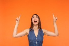 Wow Look There, Advertising Area! Surprised Happy Brunette Girl In Dress Looking Astonished And Pointing Up Copy Space On Orange Background, Showing Empty Place Above Head For Promotion. Studio Shot