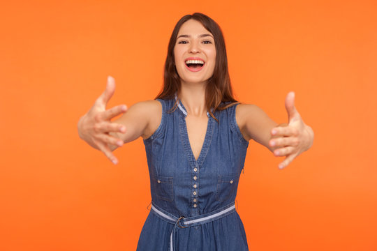 Let me hug you! Friendly brunette woman in denim dress holding arms outstretched to embrace, welcoming with hospitable expression, caring person. indoor studio shot isolated on orange background