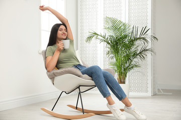 Poster - Young woman with cup of drink relaxing in rocking  chair at home