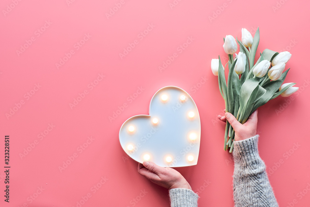 Springtime flat lay on pink, female woman hands holding bunch of white tulips, Spring flowers. Lightboard in heart shape in hands. Flat lay, top view, Mothers day or Valentine. - obrazy, fototapety, plakaty 
