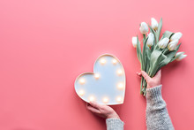 Springtime Flat Lay On Pink, Female Woman Hands Holding Bunch Of White Tulips, Spring Flowers. Lightboard In Heart Shape In Hands. Flat Lay, Top View, Mothers Day Or Valentine.