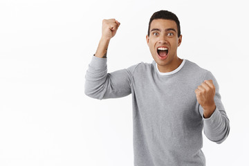 Excited, upbeat and rejoicing handsome athletic man chanting, lifting hand up in hooray, victory gesture, rooting for favorite team playing match, watching sport tv, standing white background