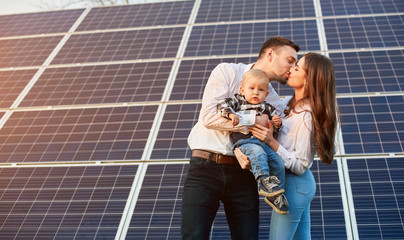 Wall Mural - Young parents are kissing in front of alternative source of energy holding their little baby boy, concept of happy parenting, copy space