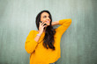 © mimagephotos - smiling young latin woman talking with cellphone and hand in hair by green wall