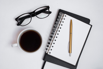 The begining of the work day. Morning at the office. Student workplace. Cup of coffee, black notebook, pen, white sheet with empty place for text and glasses on a white background. View from above.