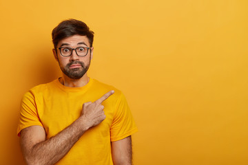 Impressed surprised man points away on blank space, shows unexpected sale or discount, wears glasses and t shirt, isolated on yellow background, shows direction or way. Advertisement concept