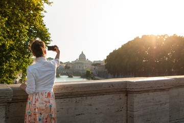 Young woman looking at St Peter's basilica from a bridge on tiber river at sunet, taking pictures with smartphone. Panoramic view cityscape of Rome, Italy.