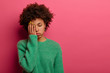Horizontal shot of exhausted dark skinned woman covers half of face, feels tired after working all night, keeps eyes closed, wears warm green sweater, isolated on pink wall with blank space for text