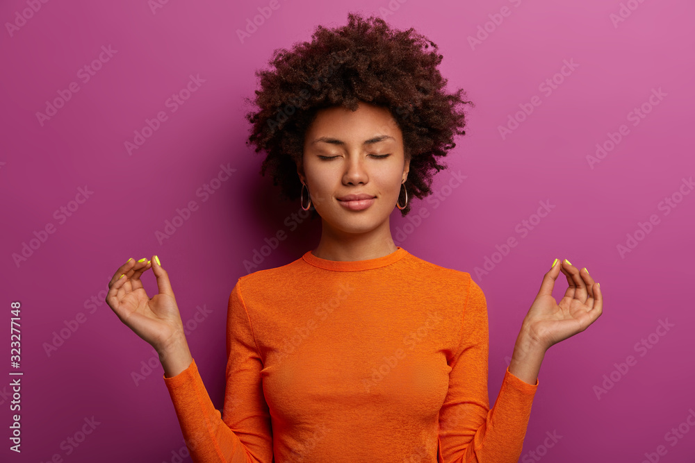 Peaceful determined curly young Afro American woman makes zen gesture, has yoga breathing practice, meditates indoor, closes eyes and wears orange jumper, isolated over vibrant purple background - obrazy, fototapety, plakaty 