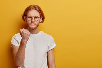 Wall Mural - Portrait of serious angry European man shows clenched fist at camera, promises to revenge, warns you about punishment, wears casual clothes, isolated over yellow background, copy space aside.