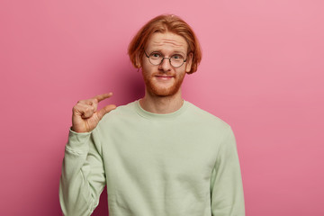 Wall Mural - Small ammount of something. Ginger bearded man claims about his poor salary, shapes something very tiny, wears glasses and jumper, tells he needs not much, isolated over rosy pastel background