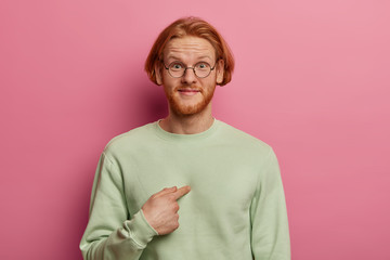 Wall Mural - You mean me? Glad bearded guy with ginger hair and beard points at himself, surprised and happy being chosen to help boss, wears green pastel jumper, isolated on rosy wall, asks about something