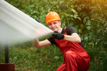 Wall Mural - Happy man worker in orange uniform and safety helmet on blurry background of solar panels and greenery. Home construction of solar station. Alternative energy sustainable concept