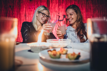 Two young women, a blonde and a redhead are cheering with glasses full of gin tonic drink and straws on a garden pary with candle on early evening