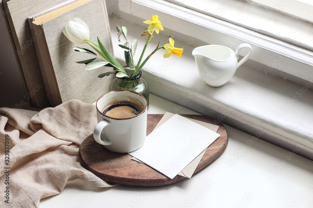 Cozy Easter spring still life. Greeting card mockup scene. Cup of coffee, books, wooden cutting board, milk pitcher and vase of flowers on windowsill. Floral composition. Yellow daffodils and tulip. - obrazy, fototapety, plakaty 