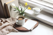 Cozy Easter Spring Still Life. Greeting Card Mockup Scene. Cup Of Coffee, Books, Wooden Cutting Board, Milk Pitcher And Vase Of Flowers On Windowsill. Floral Composition. Yellow Daffodils And Tulip.