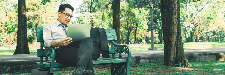 Wide panorama confident Asian businessman sitting on bench working with laptop computer at natural park, Handsome young man relaxing after work, Panoramic banner Asia business people working outside