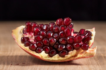 A slice of peeled pomegranate lies on a light wood kitchen board on a black background. Bright red grains of pomegranate shine beautifully. Studio photography under artificial lighting with two light 