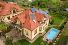 Aerial View Of A New Autonomous House With Solar Panels And Water Heating Radiators On The Roof And Green Yard With Blue Swimming Pool.