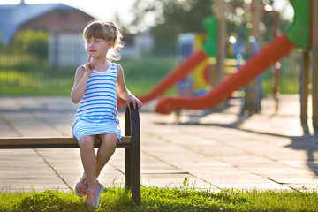 Canvas Print - Cute young girl in short dress sitting alone outdoors on playground bench on sunny summer day.