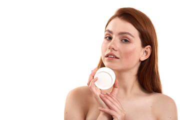 Taking care of her sensitive skin. Young attractive red-haired girl with freckles demonstrating cosmetic product and looking at camera while standing in studio against white background